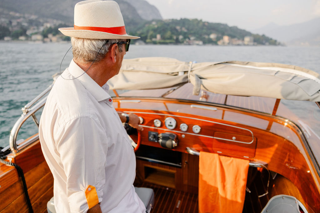 Heiratsantrag am Comer See: So wird der wichtigste Moment deines Lebens unvergesslich 7 Ein Mann mit Hut steuert ein Holzboot auf einem italienischen See, mit Hügeln und Häusern im Hintergrund - das fängt den ruhigen Charme ein, den Hochzeitsplaner wie Julia Weiß oft suchen.