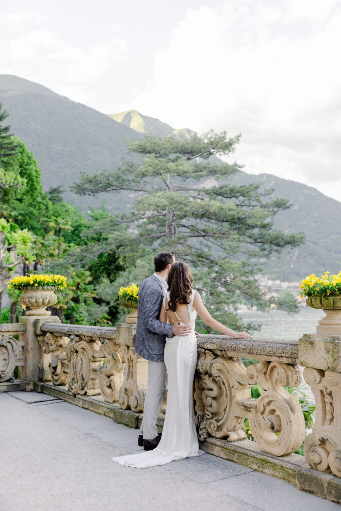 Hochzeitslocations am Comer See: 10 Luxus-Villen & Luxushotels mit direktem Seeblick am Comer See 3 Ein formell gekleidetes Paar steht auf einer Steinterrasse in Italien, mit Blick auf einen See und grüne Berge - eine Traumhochzeit, die von einem erfahrenen Hochzeitsplaner zum Leben erweckt wird.