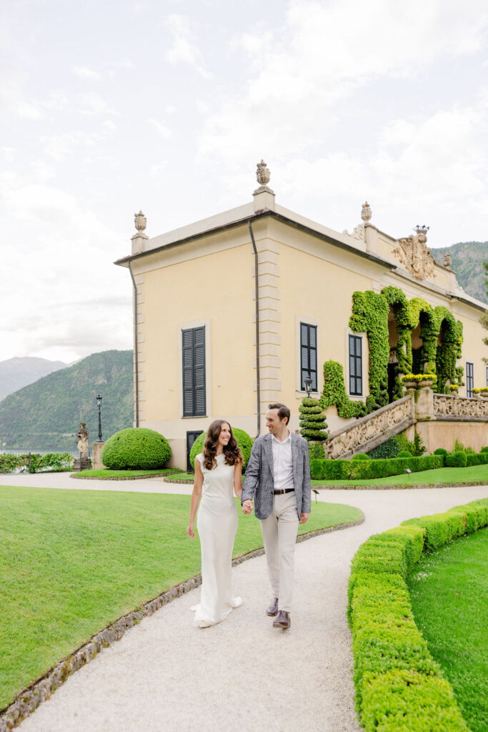 Hochzeitslocations am Comer See: 10 Luxus-Villen & Luxushotels mit direktem Seeblick am Comer See 6 Ein elegant gekleidetes Paar spaziert über einen Gartenweg an einer prächtigen Villa mit gepflegten Hecken und Blick auf die Berge vorbei - eine ideale Kulisse für eine Hochzeit in Italien, die von einem erfahrenen Hochzeitsplaner geplant wurde.