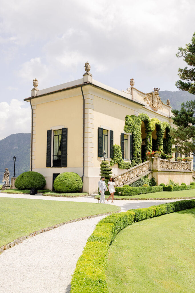 Hochzeitslocations am Comer See: 10 Luxus-Villen & Luxushotels mit direktem Seeblick am Comer See 1 Zwei Personen schlendern unter der italienischen Sonne an einer gelben Villa mit Efeu und gepflegten Gärten vorbei und fangen den Charme Apuliens ein.