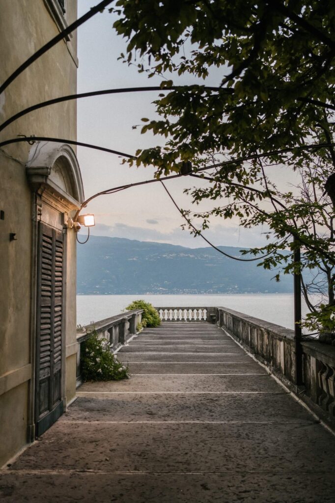 Steinweg mit Geländer mit Blick auf den ruhigen Comer See und die fernen Berge, neben einem Gebäude in der Abenddämmerung in Italien.