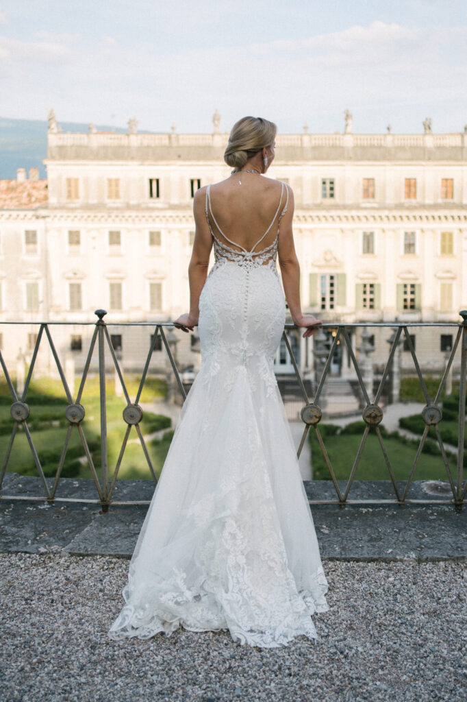 Die Braut in einem weißen Spitzenkleid steht auf einer Terrasse in Apulien vor einem eleganten historischen Gebäude, wunderschön fotografiert von Julia Weiß.