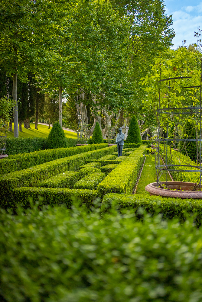 Villa Miani Rom: Heiraten in der Hochzeitslocation mit Blick über Rom 8 Gepflegter Garten mit beschnittenen Hecken, einer Statue und hohen grünen Bäumen im Hintergrund unter blauem Himmel.