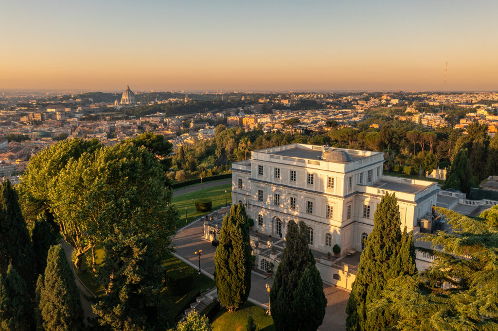 Villa Miani Rom: Heiraten in der Hochzeitslocation mit Blick über Rom 4 Eine weiße, von Bäumen umgebene Villa überblickt bei Sonnenuntergang eine Stadt unter einem klaren Himmel.