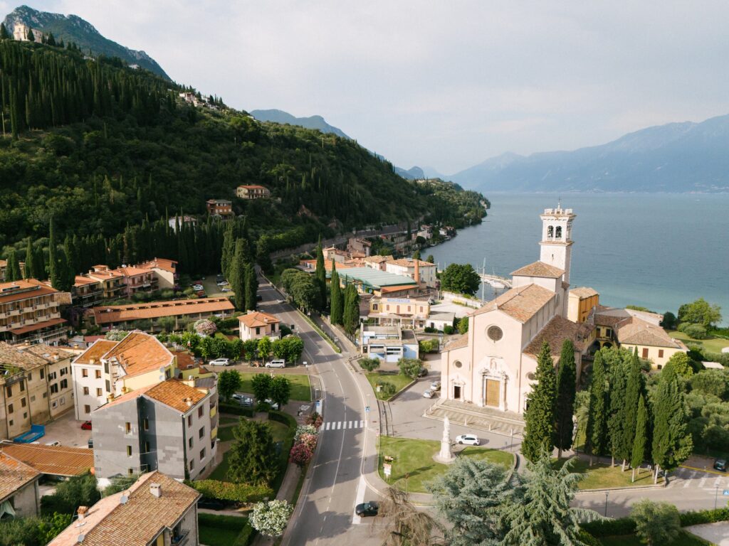 Heiraten am Gardasee, Italien - Alle Infos, Kosten & Hochzeitslocations 4 Luftaufnahme eines Dorfes am See in Italien mit einer Kirche, rot gedeckten Gebäuden und grünen Hügeln - ein zauberhafter Ort für eine von einem erfahrenen Hochzeitsplaner geplante Destination Wedding.