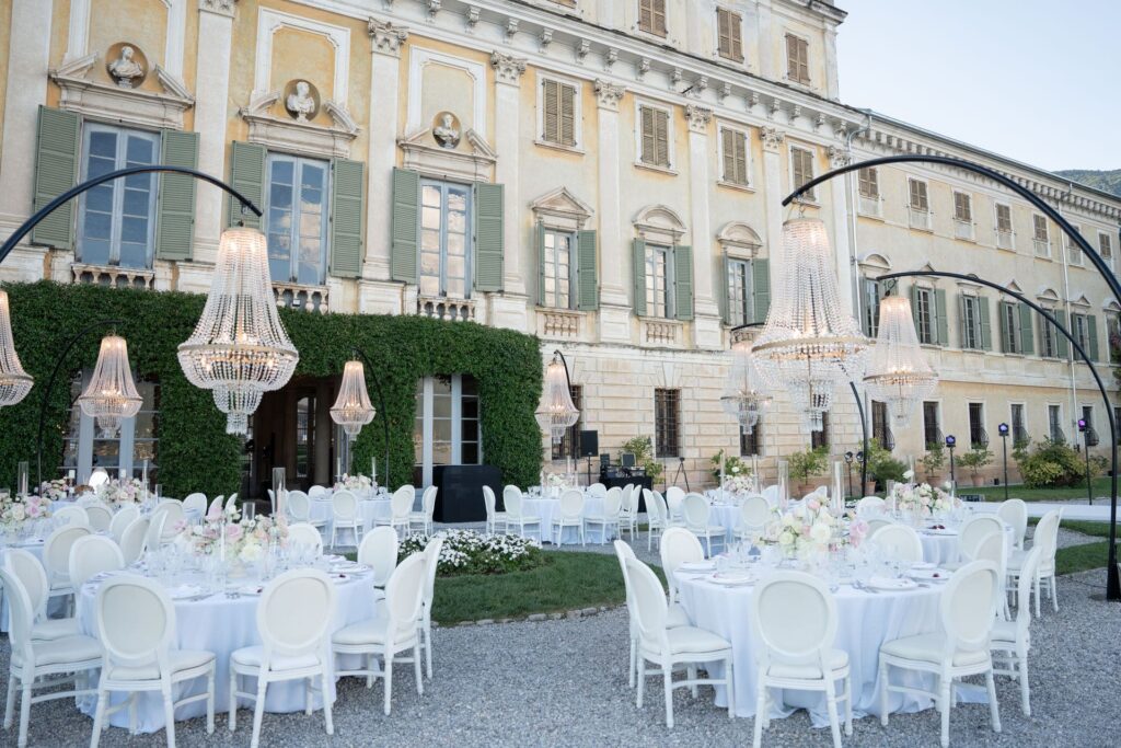 Heiraten am Gardasee, Italien - Alle Infos, Kosten & Hochzeitslocations 9 Elegante Hochzeitsfeier im Freien mit runden Tischen, weißen Stühlen und Kronleuchtern vor einem großen Gebäude - fachmännisch koordiniert von Hochzeitsplanerin Julia Weiß am wunderschönen Comer See.